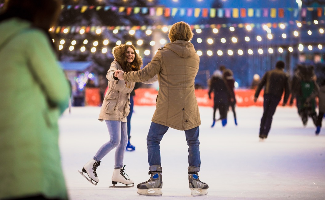 A couple ice skating on an outdoor public rink with lots of lights